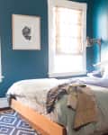 Teal bedroom with wooden bed, layered bedding, abstract wall art, and patterned rug.