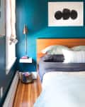 Teal bedroom with wooden headboard, abstract art, and a wall-mounted lamp. White shelf holds books and a woven basket below.