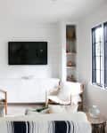 Living room with wall-mounted TV, two wooden chairs with white cushions, built-in shelves, and large windows.