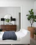 White chaise lounge with pillows and throw, facing a wooden dresser with a round mirror, flanked by black lamps and a potted plant.