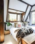 Cozy bedroom with wooden beams, large windows, a bed with decorative pillows, a fur throw, and a vintage lamp.