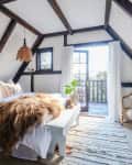 Cozy attic bedroom with exposed beams, a bed with fur throw, wicker chair, and French doors leading to a balcony.