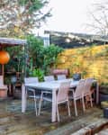 Outdoor patio with wooden deck, dining table, chairs, yellow wall, plants, and two orange lanterns hanging in a cozy seating area.