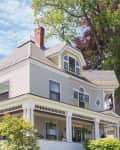 Victorian-style house with gray siding, large porch, and decorative windows, surrounded by lush greenery and trees.