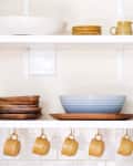 Open kitchen shelves with yellow mugs, wooden plates, blue and yellow pitchers, and a hanging plant.
