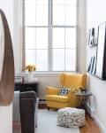 Yellow armchair with patterned cushion, pouf, and side table in a bright room with large window and wall art.