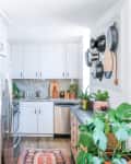 Bright kitchen with white cabinets, hanging pots, plants, and open shelving displaying mugs and decor.