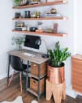 Home office with wooden desk, black chair, computer, and shelves displaying books, plants, and decorative items.