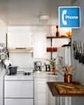 Small kitchen with white cabinets, brick wall, hanging utensils, knife rack, and a blue phone sign.