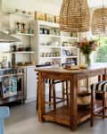 Bright kitchen with wooden island, striped chairs, wicker pendant lights, and open shelves displaying colorful dishes.