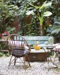 Wicker chair and table in a garden with lush greenery, a green bench, and potted plants.