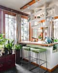 Sunlit kitchen with green plants, wooden accents, bar stools, and a blue kettle on the counter.