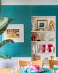 Dining room with teal walls, large plant, colorful art, and shelves displaying eclectic decor and gold accents.