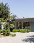 Mid-century modern house with a flat roof, surrounded by lush greenery and a gravel pathway.