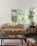 Mid-century living room with a beige sofa, fur throw, wooden side tables, potted plant, and large windows overlooking greenery.