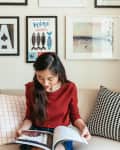 Woman in red sweater reading a magazine on a beige sofa, surrounded by patterned cushions and framed wall art.