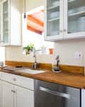 Modern kitchen with stainless steel fridge, wooden countertops, white cabinets, and potted plants by the window.
