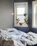 Cozy bedroom with unmade bed, gray walls, hanging bulb, potted plants on windowsill, and a small side table with a candle.