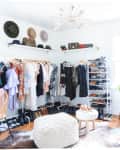 Bright walk-in closet with hanging clothes, shoe racks, hats, and bags on shelves, featuring a white pouf and wooden floor.