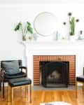 Living room with brick fireplace, blue sofa, wooden chairs, and plants on mantel. Mid-century modern decor and hardwood floor.