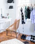 Vanity table with makeup, wicker chair, clothes rack with dresses, and sneakers on wooden floor in a bright room.