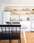 Modern kitchen with open shelves, white cabinets, black chairs, and a wooden dining table.