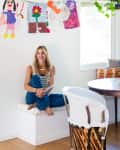 Woman sitting in a bright dining nook with a round table, colorful cushions, and children's artwork on the wall.