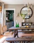 Dining room with wooden table, wicker chairs, blue doors, and a round mirror above a sideboard with decor items.