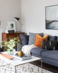 Modern living room with gray sofa, orange and black pillows, white coffee table, and fireplace. Large wall art and plants.