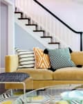 Beige sofa with geometric patterned cushions in a living room, near a staircase with black railing.