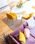 Bright living room with pink sofa, yellow chairs, round table, floral centerpiece, and a bar cart on wooden floor.