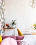 Bright living room with pink sofa, cactus wallpaper, round table with floral centerpiece, and gold chairs.