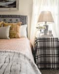 Cozy bedroom with patterned bedspread, decorative pillows, plaid-covered side table, lamp, and framed seascape painting.