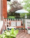 Brick patio with wooden bench, white chair, potted plants, and a blue umbrella, surrounded by lush greenery.
