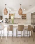 Modern kitchen with island, four bar stools, wicker pendant lights, stainless steel appliances, and white cabinetry.
