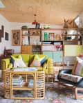 Colorful living room with rattan furniture, floral rug, and vibrant cushions, featuring a wooden shelf with eclectic decor.