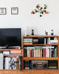 Living room with a TV on a wooden stand, bookshelves filled with books, stacked stools, and wall art.