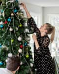 Woman decorating a Christmas tree with ornaments and garlands, while a man assists, in a bright living room.