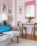 Living room with a blue sofa, colorful wall art, a pink-framed mirror, and a dining table with flowers and two chairs.