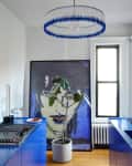 Modern kitchen with blue cabinets, abstract art, potted plant, and circular blue chandelier.