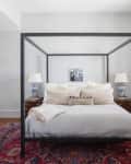 Canopy bed with white bedding, decorative pillows, blue lamps, and wooden nightstands on a patterned red rug.
