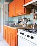 Narrow kitchen with orange cabinets, striped walls, hanging utensils, and a mirror door reflecting a hallway.