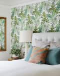 Bedroom with tropical leaf wallpaper, white bed, colorful pillows, and a wooden-framed mirror.