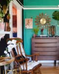 Eclectic room with green walls, wooden dresser, rocking chair, plants, and decorative wall art.