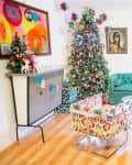 Festive living room with a decorated Christmas tree, teal sofa, colorful chairs, and vibrant wall art.