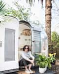 Woman sitting at the entrance of a vintage trailer with potted plants and palm trees in the background.