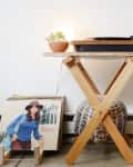 Turntable on wooden stand with a potted plant, next to a stack of vinyl records featuring a woman in a hat.