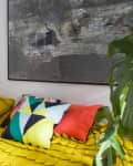Yellow bed with colorful geometric pillows, large abstract wall art, and a green monstera plant.