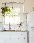 Bright kitchen with white cabinets, granite countertop, sink, hanging plants, and a bowl of colorful fruits.