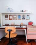 Home office with wooden chair, desk, pink tool chest, clipboards on wall, and shelf with vases and decor items.